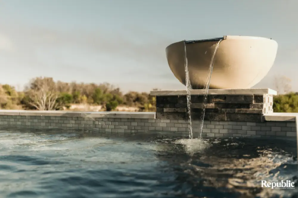 Aerial view of a modern, two-section swimming pool with crystal-clear water, featuring a raised hot tub area. Surrounding concrete and grass